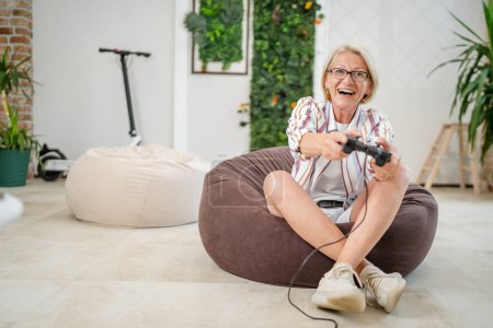 Téléchargez les photos : Une femme blonde caucasienne mature avec des lunettes jouer console de jeu vidéo en utilisant un contrôleur joystick tout en étant assis à la maison personnes réelles loisirs concept copier espace heureux sourire célébrer gagner de l'espace de copie - en image libre de droit