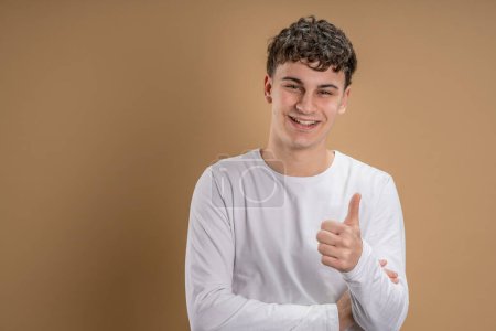 Photo for Portrait of one caucasian man 20 years old looking to the camera in front of almond color studio background smiling wearing casual shirt thumb up copy space - Royalty Free Image