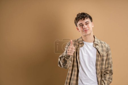 Téléchargez les photos : Portrait d'un homme caucasien de 20 ans regardant vers l'appareil photo en face de couleur amande studio arrière-plan souriant portant chemise décontractée pouce vers le haut de l'espace de copie - en image libre de droit