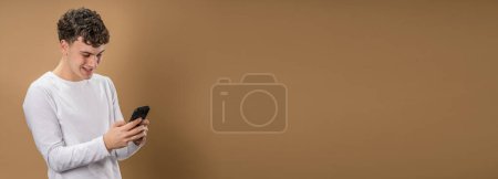 Téléchargez les photos : Un homme caucasien adolescent homme garçon utiliser smartphone téléphone mobile pour la navigation sur Internet en ligne réseau social ou sms messages texte studio shot beige fond heureux sourire - en image libre de droit