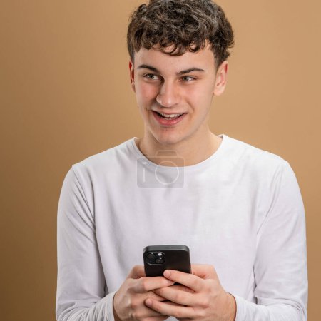 Foto de Un hombre caucásico varón adolescente uso smartphone teléfono móvil para la navegación en línea de Internet red social o SMS mensajes de texto estudio disparo beige fondo sonrisa feliz - Imagen libre de derechos