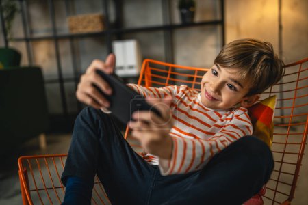 Téléchargez les photos : Un garçon assis à la maison tenir téléphone mobile smartphone jouer à des jeux vidéo - en image libre de droit