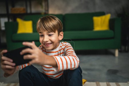 Téléchargez les photos : Un garçon assis à la maison tenir téléphone mobile smartphone jouer à des jeux vidéo - en image libre de droit