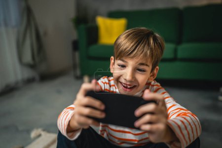 Foto de Un niño se sienta en casa celebrar teléfono móvil smartphone jugar videojuegos - Imagen libre de derechos