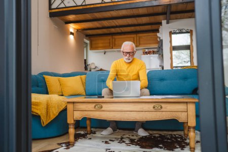 Téléchargez les photos : Un homme caucasien senior grand-père avec barbe et lunettes de travail sur ordinateur portable utiliser l'ordinateur à la maison - en image libre de droit
