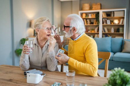 Photo for Senior couple woman take medicine or supplement vitamin treatment cure while her husband old caucasian man sit beside at the table at home real people family old people retirement and health concept - Royalty Free Image