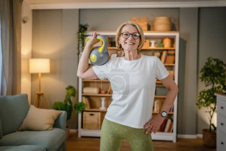 One mature caucasian blonde woman training with kettlebell girya russian bell at home female exercise in her apartment healthy lifestyle concept