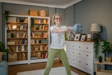 One mature caucasian blonde woman training with kettlebell girya russian bell at home female exercise in her apartment healthy lifestyle concept