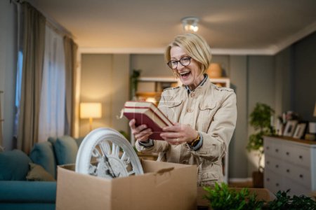 Photo for One senior woman pensioner grandmother moving in new apartment taking her stuff belongings and plants in or out of boxes packing or unpacking real person hold books copy space - Royalty Free Image