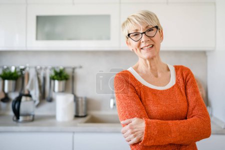 Foto de Primer plano retrato de una mujer mayor con el pelo corto sonrisa feliz emoción positiva copia espacio de pie en casa en la cocina gafas de desgaste interior - Imagen libre de derechos