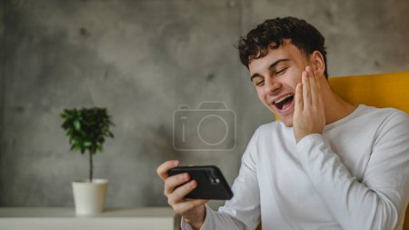 Téléchargez les photos : Portrait de garçon adolescent assis à la maison utiliser smartphone pour regarder en ligne podcast vidéo à la maison - en image libre de droit