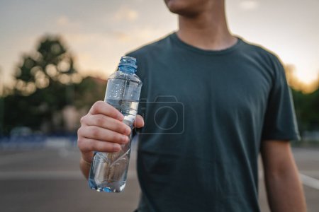 Téléchargez les photos : Homme caucasien adolescent ouvert bouteille en plastique d'eau en plein air dans une boisson ensoleillée tout en tenant basket-ball - en image libre de droit