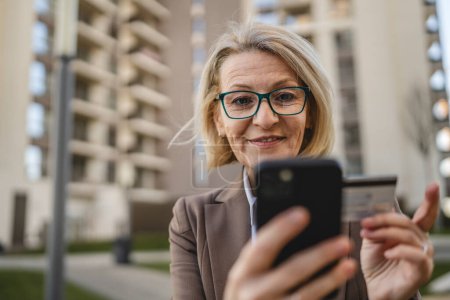 Téléchargez les photos : Une femme est assise devant un immeuble, regardant un téléphone portable, tenant une carte de crédit et effectuant un paiement en ligne - en image libre de droit