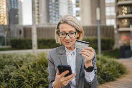 Photo for One woman is sitting in front of a building looking at a mobile phone and holding a credit card and making an online payment - Royalty Free Image