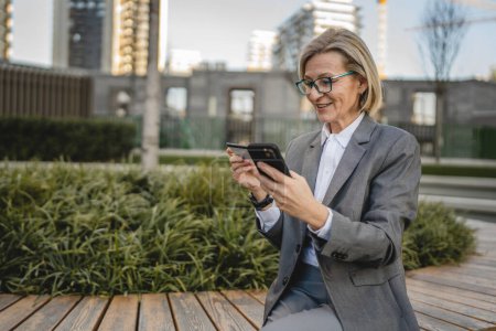 Foto de Una mujer está sentada frente a un edificio mirando un teléfono móvil y sosteniendo una tarjeta de crédito y haciendo un pago en línea - Imagen libre de derechos