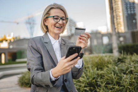 Téléchargez les photos : Une femme est assise devant un immeuble, regardant un téléphone portable, tenant une carte de crédit et effectuant un paiement en ligne - en image libre de droit