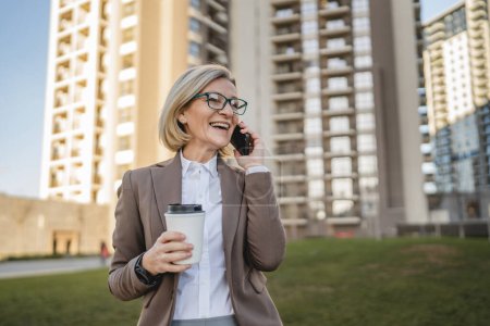 Foto de Una mujer madura al aire libre frente a edificios hablando por teléfono móvil y sosteniendo un café para llevar, negocio moderno y concepto de estilo de vida - Imagen libre de derechos