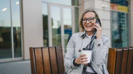 Téléchargez les photos : Une femme mûre à l'extérieur devant des bâtiments parlant sur un téléphone portable et tenant un café à emporter, concept moderne d'affaires et de style de vie - en image libre de droit