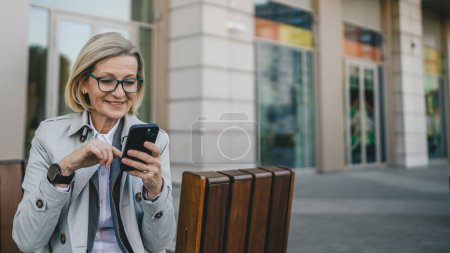 Foto de Una mujer moderna madura hembra de pie en la ciudad de uso al aire libre teléfono móvil enviar mensajes de texto SMS o navegar por Internet en línea rubia hembra con gafas espacio de copia - Imagen libre de derechos