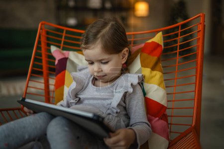 Téléchargez les photos : Une fille s'asseoir à la maison tenir tablette numérique regarder et jouer à des jeux vidéo - en image libre de droit