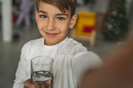 Téléchargez les photos : Un garçon caucasien enfant se tenir à la maison tenir verre d'eau soif boisson vue de face portrait porter chemise blanche regarder tot il appareil photo copier espace heureux sourire - en image libre de droit