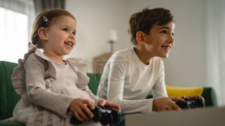 Photo for Two children small caucasian brother and sister happy children siblings boy and girl playing video game console using joystick or controller while sitting at home real people family leisure concept - Royalty Free Image