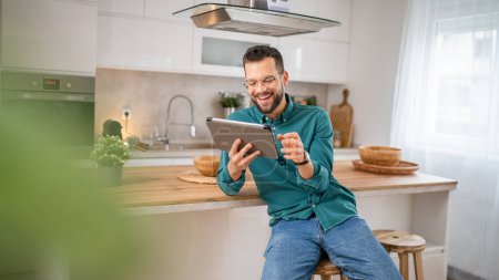 Téléchargez les photos : Un homme adulte caucasien mâle tenir un travail tablette numérique ou naviguer sur Internet sourire heureux à la maison assis confiant quotidien matin routine copie espace personne réelle - en image libre de droit