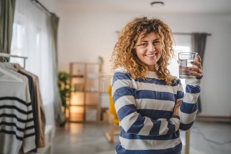 Téléchargez les photos : Une femme belle femme adulte à la maison avec un verre d'eau bouclés cheveux heureux sourire mode de vie sain concept d'hydratation - en image libre de droit