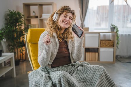 Téléchargez les photos : Une femme caucasienne adulte femelle avec écouteurs utilisent un téléphone mobile à la maison pour écouter la musique en ligne sourire heureux chanter joie avoir du plaisir seul espace de copie - en image libre de droit