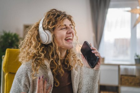 Téléchargez les photos : Une femme caucasienne adulte femelle avec écouteurs utilisent un téléphone mobile à la maison pour écouter la musique en ligne sourire heureux chanter joie avoir du plaisir seul espace de copie - en image libre de droit