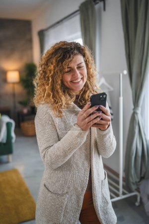 Foto de Una mujer caucásica mujer adulta con el pelo rizado en el hogar usar teléfono móvil SMS SMS mensajes de texto o navegar por Internet en línea red social - Imagen libre de derechos