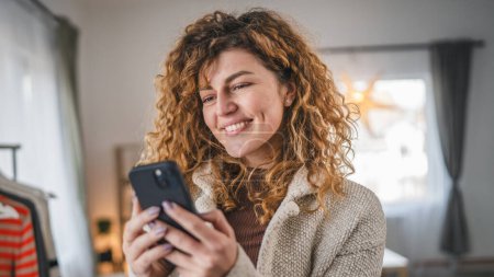 Foto de Una mujer caucásica mujer adulta con el pelo rizado en el hogar usar teléfono móvil SMS SMS mensajes de texto o navegar por Internet en línea red social - Imagen libre de derechos