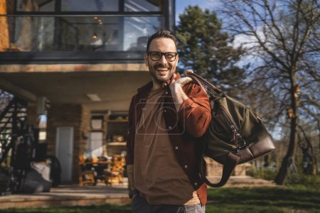 Téléchargez les photos : Portrait d'un homme heureux homme caucasien avec sac de voyage à sa maison - en image libre de droit