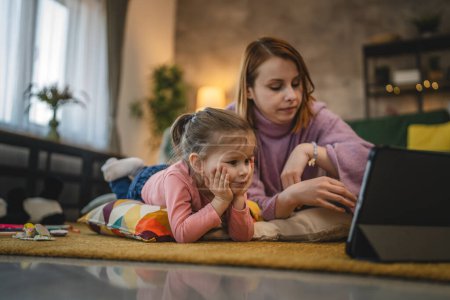 Téléchargez les photos : Mère et fille utilisent une tablette numérique pour regarder une vidéo en ligne à la maison - en image libre de droit