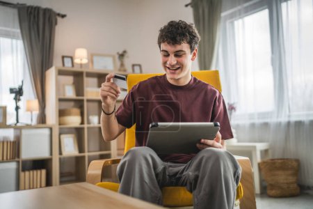 Téléchargez les photos : Un adolescent jeune homme caucasien homme assis à la maison utiliser carte de crédit achats en ligne utiliser Internet pour l'achat en ligne active moderne e-banking - en image libre de droit