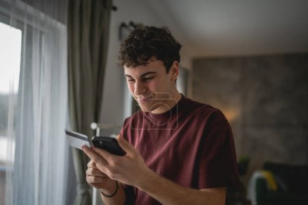 Téléchargez les photos : Un adolescent jeune homme caucasien homme assis à la maison utiliser carte de crédit achats en ligne utiliser Internet pour l'achat en ligne active moderne e-banking - en image libre de droit