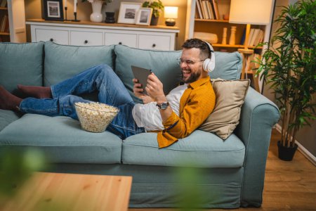 Téléchargez les photos : Un homme adulte caucaïen mâle sur le canapé-lit à la maison regarder le film vidéo en ligne utiliser une tablette numérique ou avoir un appel vidéo - en image libre de droit