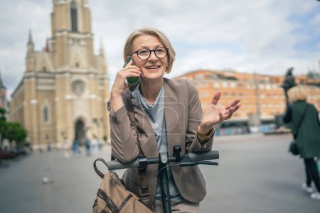 Photo for One woman mature caucasian blonde female stand outdoor with electric kick push scooter use mobile phone smartphone to make a call talk in the city stand alone confident real people copy space - Royalty Free Image