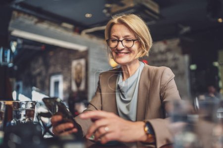 Photo for One stylish mature blonde Caucasian woman wearing eyeglasses sit comfortably in a cafe texting and browsing the internet on her smartphone modern lifestyle digital interactions of mature individuals - Royalty Free Image