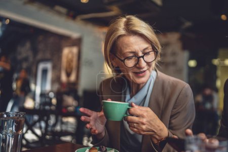 Photo for Mature caucasian woman sit in serene ambiance of a cozy dark cafe With a cup of coffee in hand wear eyeglasses enjoy the moment real person copy space - Royalty Free Image