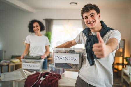 Photo for Mother and son stand at home choose clothes for donation teenage mad and caucasian woman sorting wardrobe to the boxes in their apartment living room - Royalty Free Image