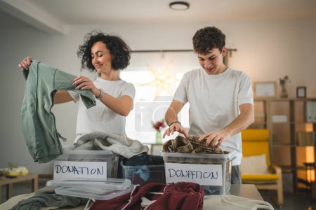 Photo for Mother and son stand at home choose clothes for donation teenage mad and caucasian woman sorting wardrobe to the boxes in their apartment living room - Royalty Free Image
