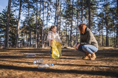 Téléchargez les photos : Mère et fille petite fille caucasienne tout-petit et femme collectent les déchets de bouteilles en plastique du parc naturel forestier le mettent dans le sac - en image libre de droit