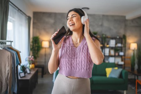 Foto de Una mujer caucásica mujer adulta con auriculares usar el teléfono móvil en casa para escuchar la música en línea sonrisa feliz cantar alegría divertirse solo - Imagen libre de derechos
