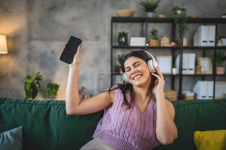 Foto de Una mujer caucásica mujer adulta con auriculares usar el teléfono móvil en casa para escuchar la música en línea sonrisa feliz cantar alegría divertirse solo - Imagen libre de derechos