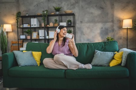 Téléchargez les photos : Une femme caucasienne adulte femelle avec casque utiliser téléphone mobile à la maison pour écouter la musique en ligne sourire heureux chanter joie avoir du plaisir seul - en image libre de droit