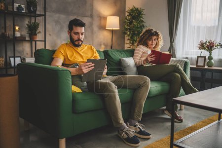 Foto de Hombre y mujer caucásico pareja adulta leer libro y utilizar tableta digital en casa en sofá cama separado - Imagen libre de derechos