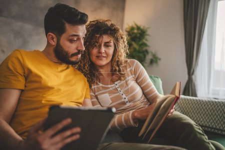 Foto de Hombre y mujer caucásico pareja de adultos leer libro y utilizar la tableta digital en casa en el sofá cama - Imagen libre de derechos