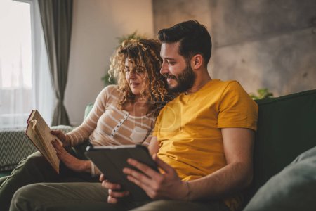 Photo for Man and woman caucasian adult couple read book and use digital tablet at home on sofa bed - Royalty Free Image