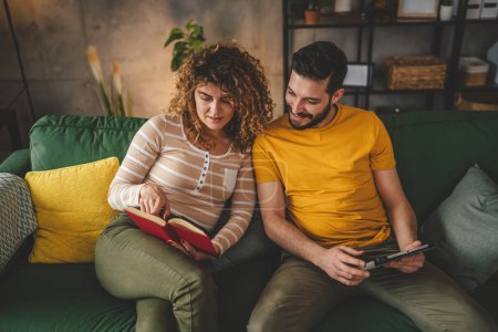 Téléchargez les photos : Homme et femme caucasien adulte couple lire livre et utiliser tablette numérique à la maison sur canapé-lit - en image libre de droit